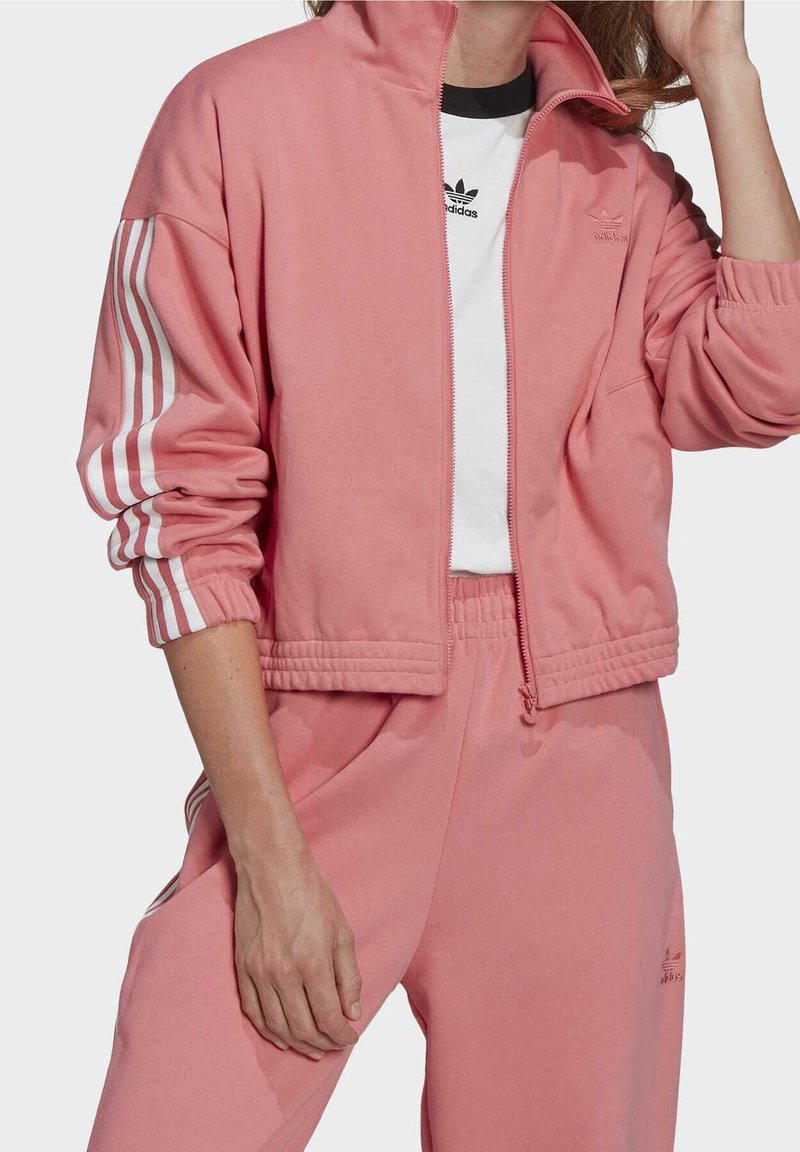 Rosa zip-up jacka med vita ränder på ärmarna, i kombination med matchande byxor och en svart-vit logotyp T-shirt under. Mjuk tygstruktur.