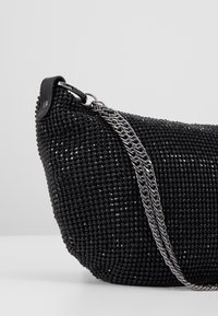 Petit sac à main noir couvert de perles brillantes avec une bandoulière chaîne argentée et un accent en cuir sur un fond gris.