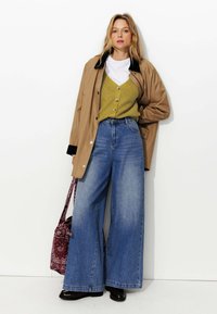 Veste imperméable beige avec accents noirs, cardigan en mailles vertes sur un t-shirt blanc, jeans en denim bleu à jambes larges, et un sac bordeaux à motifs.