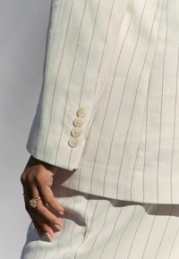 Bílý blazer s jemným proužkem a čtyřmi krémovými knoflíky na rukávu. Hladká textura látky, přiléhavý střih a diskrétní detaily podél klopy.