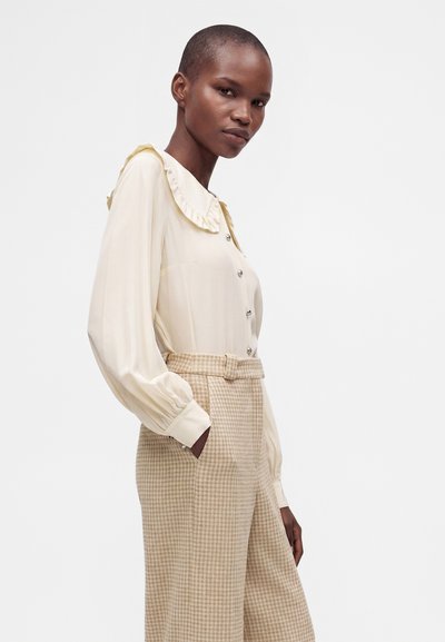 Jeune femme portant une blouse crème avec un col à volants et un pantalon beige à carreaux taille haute, posant avec une main dans la poche sur fond blanc.