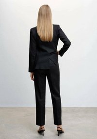 Černý oblek na míru s přiléhavým sakem a zadním rozventem a slim fit kalhotami. Doplněno černými sandály na vysokém podpatku. Hladká textilie.