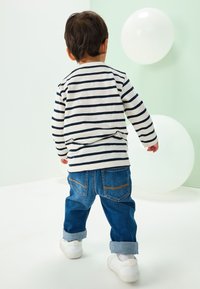 Kreemika ja mereväe triibuline pikakäise triiksärk, millel on ülesrullitud sinised teksapüksid ja valged tossud, vaadelduna tagantvalgustusega heleda tausta ees.