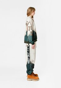 Sudadera oversized con estampado de montaña y acentos verdes, combinada con pantalones estampados que presentan un diseño en marrón y beige. Se lleva con botas color arena.