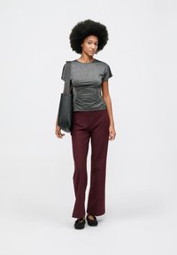 Mulher com cabelo afro, vestindo uma blusa cinza franzida, calças burgundy, sapatos pretos baixos e a segurar uma bolsa preta sobre o ombro.