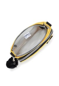 Kellomerkki, jossa on keltainen ja musta kangas, pehmeä musta pom-pom -koriste, vuorattu sisäosa ja logotunnus sisällä.