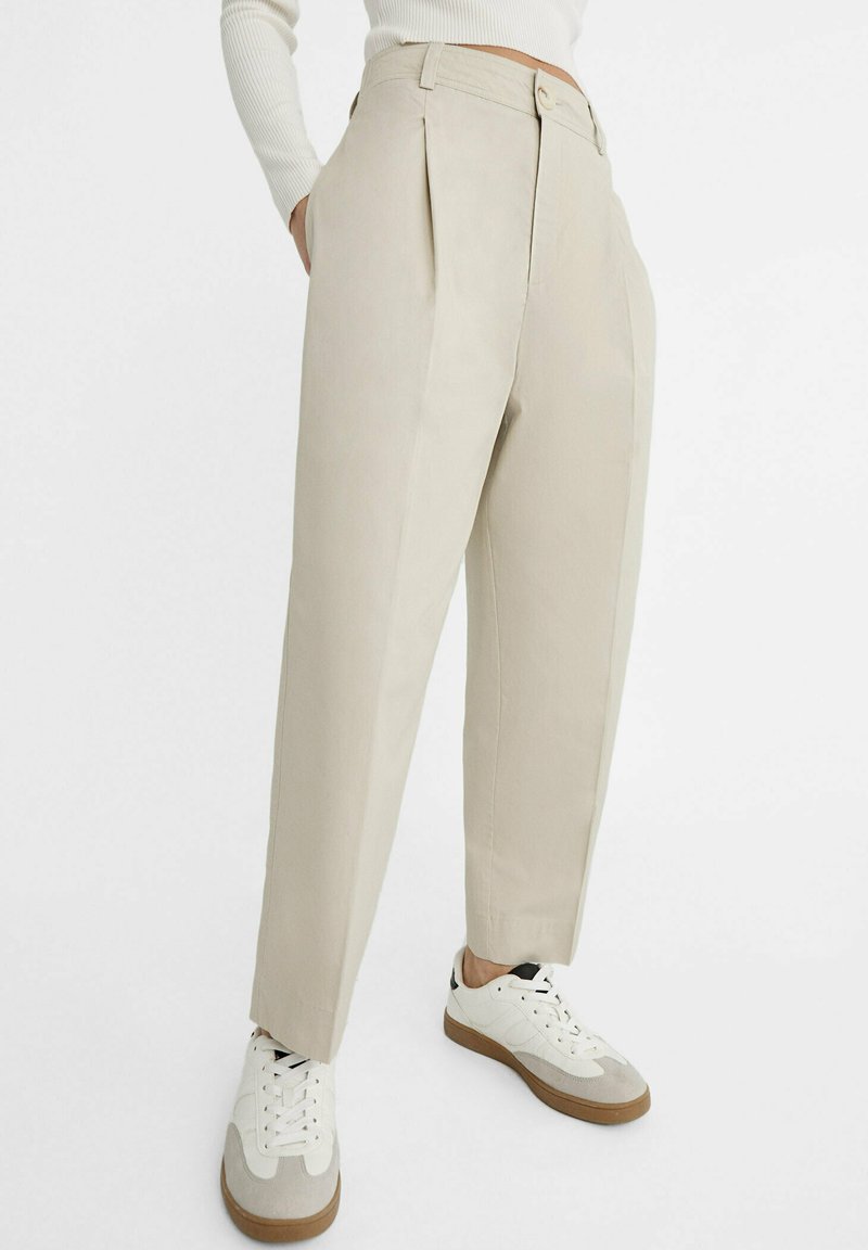 Pantaloni beige a pieghe realizzati in un tessuto leggero, con vita alta, tasche laterali e design a gamba affusolata. Indossati con sneakers bianche.