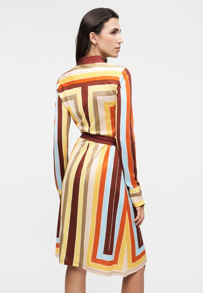 Γυναίκα με μακρυμάνικο φόρεμα μέχρι το γόνατο με κάθετες και γεωμετρικές ρίγες σε καφέ, πορτοκαλί, κίτρινο, μπλε και μπεζ, εμφανίζεται από πίσω.