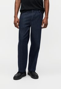 Άνδρας που φοράει σκούρο μπλε τζιν παντελόνια με λεπτομέρειες ροζ ραφές, συνδυασμένα με μαύρα παπούτσια χωρίς κορδόνια, στέκεται μπροστά σε απλό φόντο.