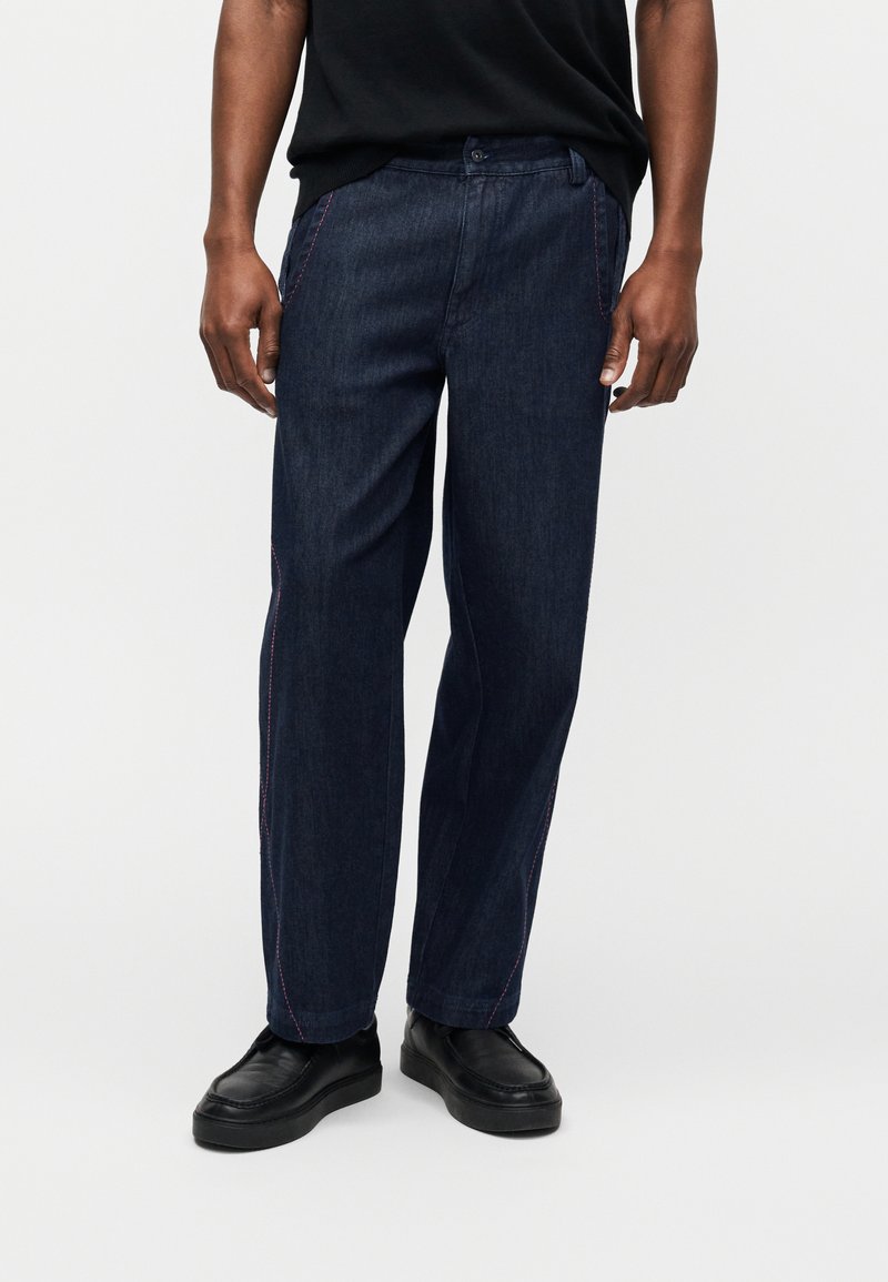 Άνδρας που φοράει σκούρο μπλε τζιν παντελόνια με λεπτομέρειες ροζ ραφές, συνδυασμένα με μαύρα παπούτσια χωρίς κορδόνια, στέκεται μπροστά σε απλό φόντο.