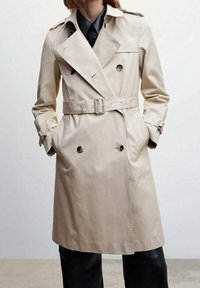 Beżowy płaszcz trench z gładkiego materiału, z podwójnym zapięciem na guziki, paskiem w talii i naramiennikami.