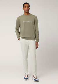 Felpa verde oliva con testo bianco in corsivo, abbinata a pantaloni tapered crema chiaro. L'outfit è completato da sneakers multicolori.