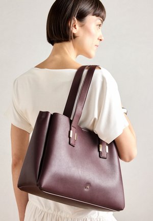 Vrouw met kort haar in een witte outfit, draagt een grote donkerbruine leren shopper over haar schouder tegen een effen achtergrond.