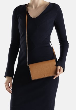 Braune Leder-Crossbody-Tasche mit strukturierter Oberfläche und langem Trageriemen, gehalten gegen ein dunkel gefärbtes Kleid mit V-Ausschnitt.