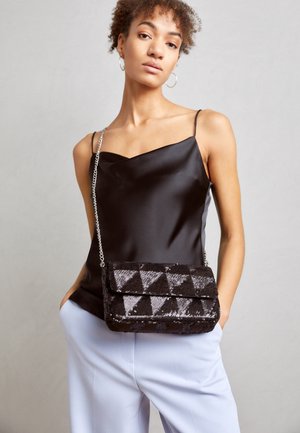 Vrouw draagt zwarte satijnen camisoleshirt, lichtgekleurde broek en een zwarte, met pailletten bezaaide crossbodytas met kettingriem, tegen een effen achtergrond.