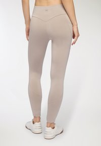 Beżowe legginsy z wyższym stanem o gładkiej, elastycznej teksturze. Posiadają skośne szwy oraz logo na tylnej części pasa, zestawione z białymi sneakersami.