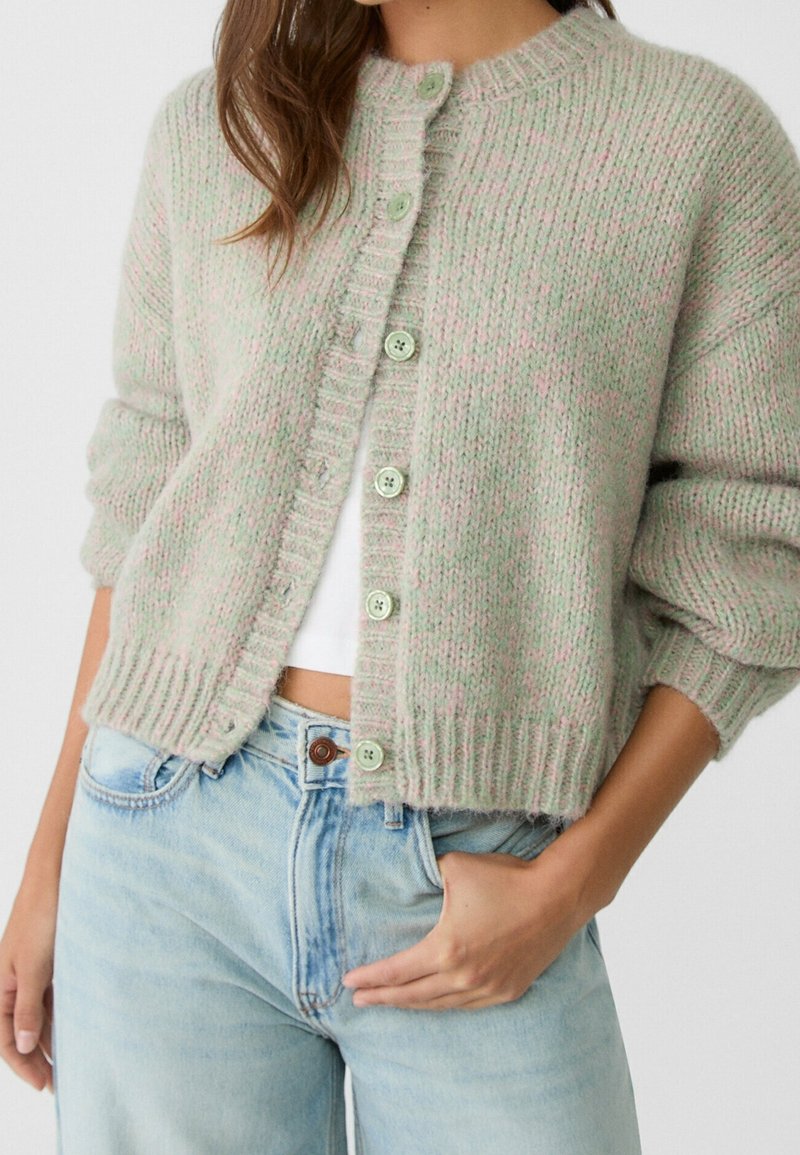 Femme portant un cardigan tricoté vert clair et rose boutonné sur un top court blanc avec un jean bleu clair, main dans la poche.
