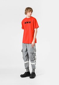 Camiseta corta roja con letras negras "MWM", combinada con pantalones cargo grises que tienen múltiples bolsillos y detalles en blanco, usados con calzado negro.