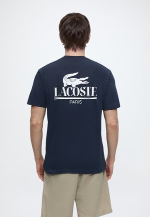 Mann trägt ein marineblaues Lacoste T-Shirt mit weißem Krokodillogo und dem Schriftzug „LACOSTE PARIS“ auf dem Rücken, kombiniert mit beigen Shorts.