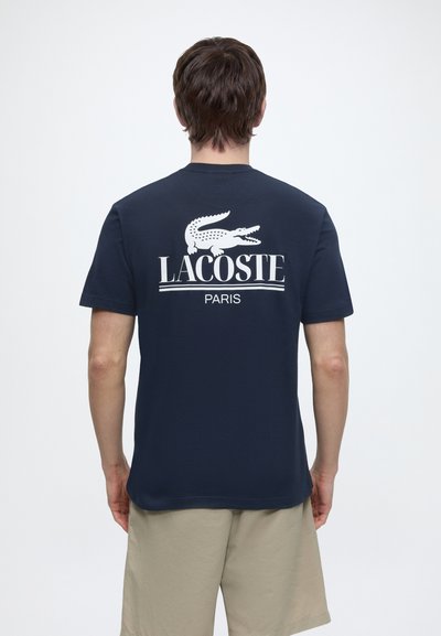 Мъж, носещ тъмносиня тениска Lacoste с бяло лого на крокодил и текст „LACOSTE PARIS“ на гърба, комбинирана с бежови шорти.