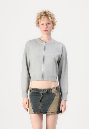 Grå kortermet genser med glidelås foran, ribbestrikket hals og ermer, kombinert med et denimskjørt med lapper og glidelåsdetaljer i variierende farger.