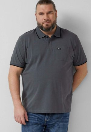 Brodaty mężczyzna z włosami związanymi do tyłu, ubrany w ciemnoszary polo i niebieskie dżinsy, stoi z jedną ręką za plecami.