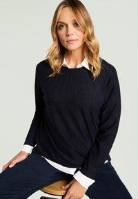 Granatowy sweter z dzianiny o prążkowanej fakturze, ozdobiony małymi aplikacjami. Noszony na białej koszuli z kołnierzykiem, z okrągłym dekoltem i długimi rękawami.