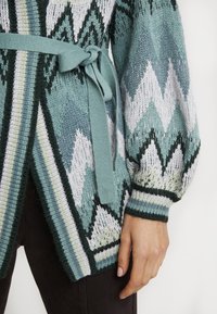 Stickad cardigan med geometriska zigzagmönster i teal, vitt och mörkgrönt, knuten i midjan, bärs med mörka byxor och neutralt nagellack.