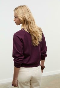 Maglione lavorato a maglia viola con polsini e orlo a coste, indossato sopra pantaloni beige chiaro a vita alta. Texture liscia, vestibilità comoda.