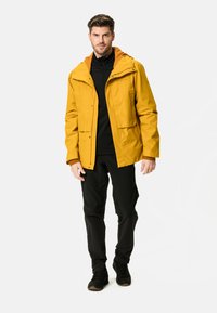Gelbe wasserdichte Jacke mit Kapuze, ausgestattet mit einem Reißverschluss vorne und zwei Fronttaschen, kombiniert mit einem schwarzen Unterziehshirt und schwarzen Hosen.