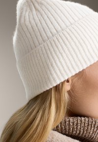 Beanie a coste bianco con punta a spirale, abbinato a un colletto a coste marrone. Texture morbida, adatta per il freddo.