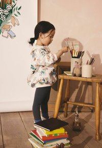 Lapsi kukkakuvioisessa svetarissa ja tummissa ribbineuloksisissa trikoissa seisoo pinottujen kirjojen päällä puisella pöydällä ja yltää värikyniä kohti.