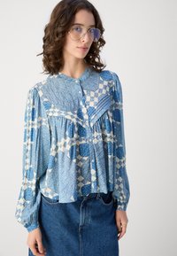Lys blå bluse med rutete og sirkulære mønstre, knapp foran og plisserte detaljer ved skuldrene; brukt med et denimskjørt.