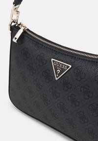 Pochette bracelet noire Guess avec motif logo embossé subtil, fermeture éclair dorée et emblème métallique triangulaire Guess USA à l'avant.
