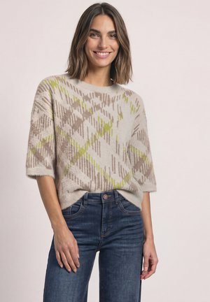 Femme aux cheveux châtain mi-longs souriante, portant un pull beige et vert à motifs à manches courtes et un jean bleu, debout devant un fond clair.