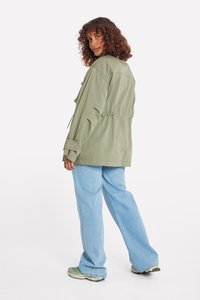Veste vert olive à coupe décontractée, taille ajustable, grandes poches et poignets à boutons, associée à un jean bleu clair à jambes larges et des baskets.