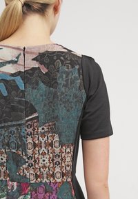 Černé šaty s krátkým rukávem, které mají multikolorovanou, vzorovanou zadní část s abstraktními designy a texturami, včetně květinových a geometrických prvků.