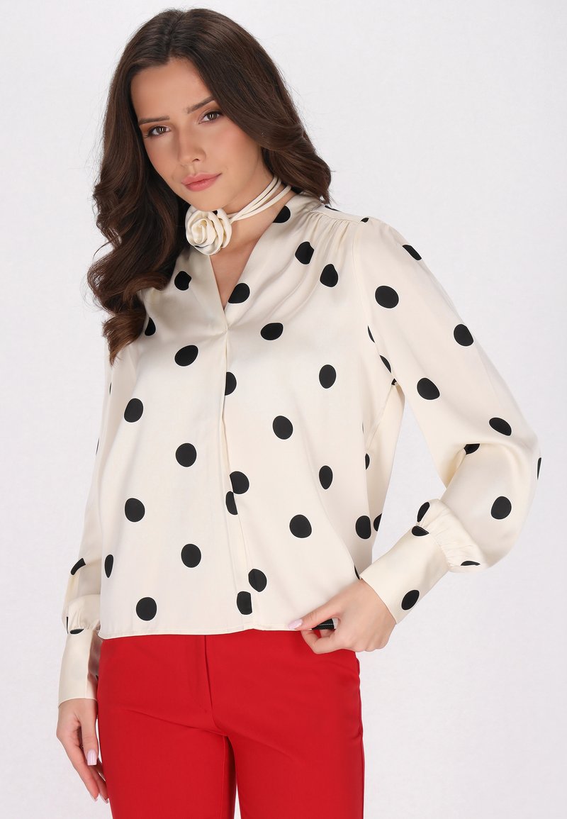 Blusa crema con pois neri, chiusura con bottoni e maniche lunghe. Abbinata a pantaloni rossi, con un dettaglio coordinato al collo.