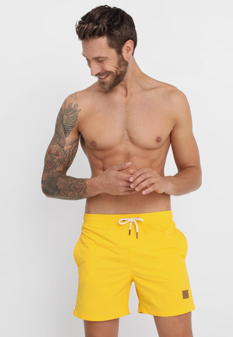 Shorts de bain jaunes vifs avec un cordon de serrage à la taille, fabriqués en matériau léger. Présente un petit patch logo marron à l'avant.