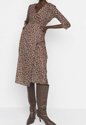 Frau trägt ein braunes Wickelkleid mit Animalprint, dreiviertel Ärmeln und kniehohen braunen Lederstiefeln mit goldener Kettendetail.