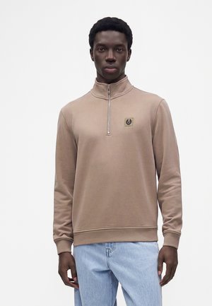 Junger Mann trägt einen beigen Viertel-Reißverschluss-Pullover mit Logo auf der Brust und hellblaue Jeans, steht vor einem schlichten weißen Hintergrund.