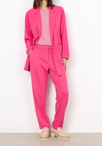 Giacca rosa brillante e pantaloni abbinati, indossati con una maglietta a righe. Il tessuto appare liscio, con una vestibilità comoda e tasche discrete.