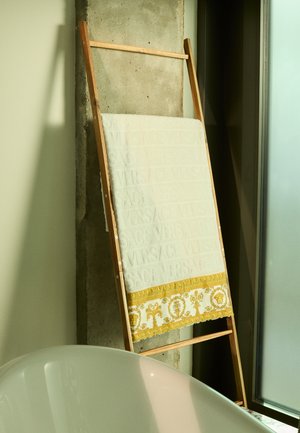 White towel with embossed "VERSACE" logo and gold baroque border, displayed on a wooden stand against a concrete wall. Bathtub partially visible.