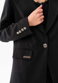 Zwarte blazer met een gladde textuur, voorzien van zilveren knopen, één borstzak en een label met de tekst "blue shadow" op de zak.