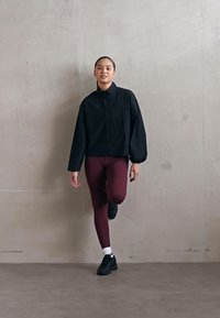 Schwarze Zip-Jacke mit hohem Kragen, kombiniert mit kastanienfarbenen Leggings und schwarzen Sneakern, vor einem neutralen Hintergrund stehend.