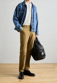 Denimjack, lichtgrijze polo, tan broek en zwarte leren schoenen. Houdt een zwarte sporttas vast, weergegeven op een houten vloer.