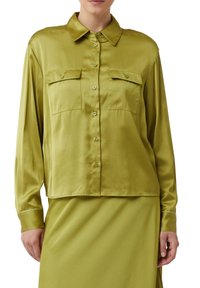 Woman wearing a long-sleeve olive green satin shirt with buttoned front and two buttoned flap chest pockets, paired with a matching skirt.