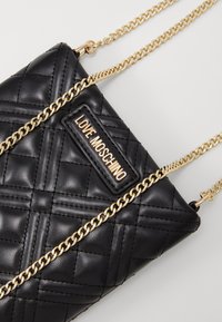 Černá prošívaná kožená kabelka přes rameno se zlatým řetízkem a emblémem "Love Moschino". Má diamantový vzor a hladký povrch.