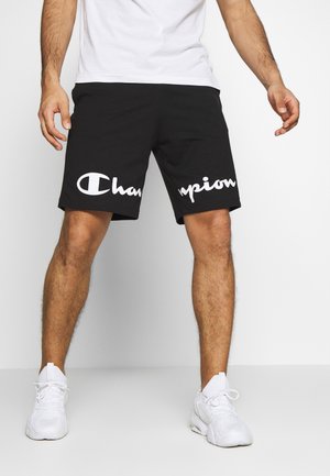 Homme portant un short noir avec le logo blanc "Champion", des baskets blanches et un t-shirt blanc, debout, jambes écartées, sur un fond uni.