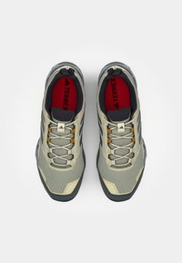 Chaussures de randonnée en mesh beige avec des semelles gris foncé, des accents orange et des détails réfléchissants. Présente une semelle intérieure rembourrée et un capo renforcé.